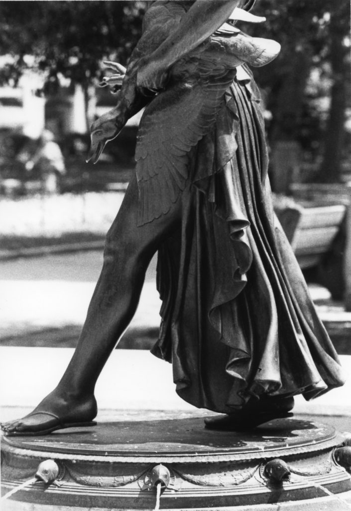 A black-and-white photo shows the lower half of a bronze statue of a girl, stepping forward with one bare leg and the other covered by a flowing draped garment. A duck-like bird is held near her hip, and she stands on a circular fountain base with small spouts.