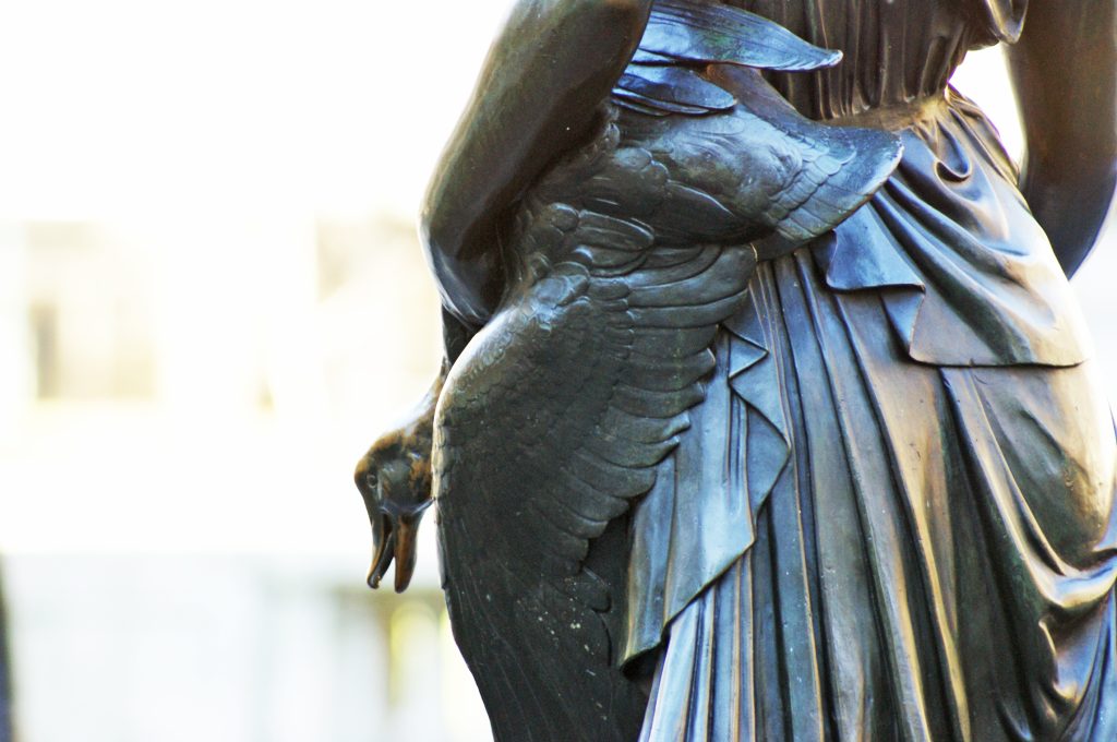 A close-up of a bronze statue shows the back of a draped figure holding a duck under one arm. The duck's head and feathered wing are visible against the figure's pleated garment.
