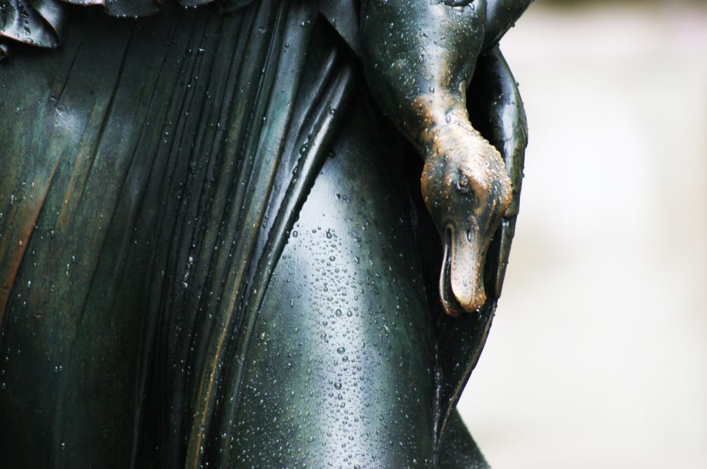 A close-up of a bronze sculpture features a duck's head emerging from draped fabric, all covered in water droplets.