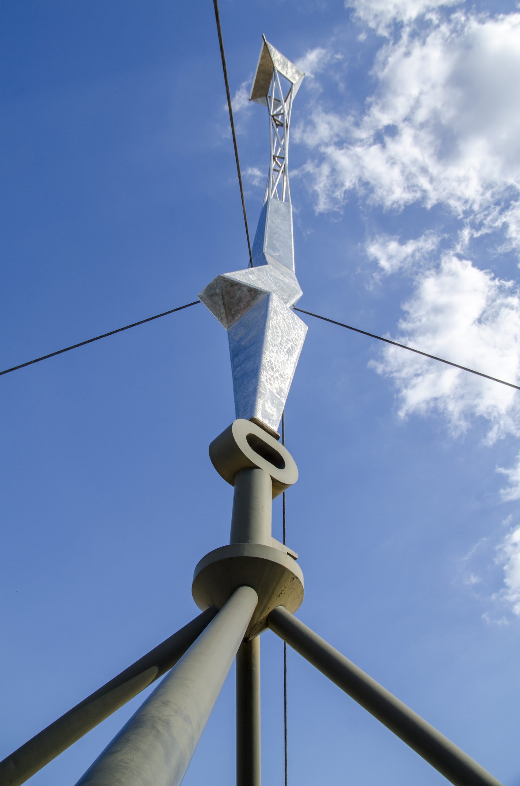 A low-angle view looking straight up a tall, abstract stainless steel tower sculpture composed of jagged geometric facets, anchored to a thick dark gray base and stabilized by multiple taut guy wires stretching into a bright blue sky with scattered clouds.