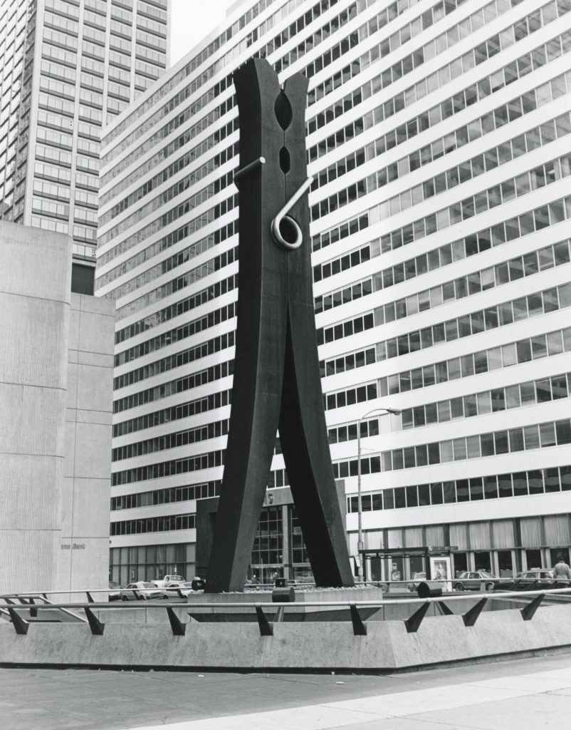 A black-and-white photograph shows a monumental, dark sculpture of a clothespin standing in an urban plaza. The two large, black legs of the clothespin meet at the top, where a silver-colored coiled spring is visible. Tall, modern buildings with numerous windows rise in the background.