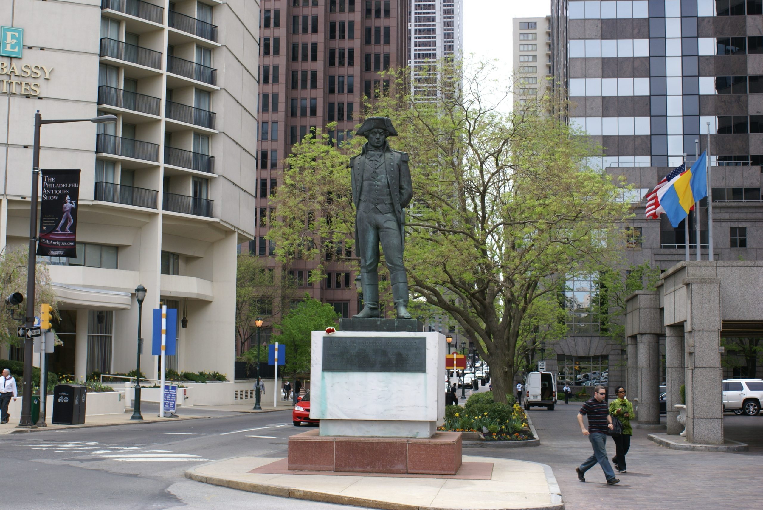 The bronze statue of General Tadeusz Kosciuszko stands on a concrete pedestal along a city street corner in Philadelphia, surrounded by high-rise buildings and green trees.