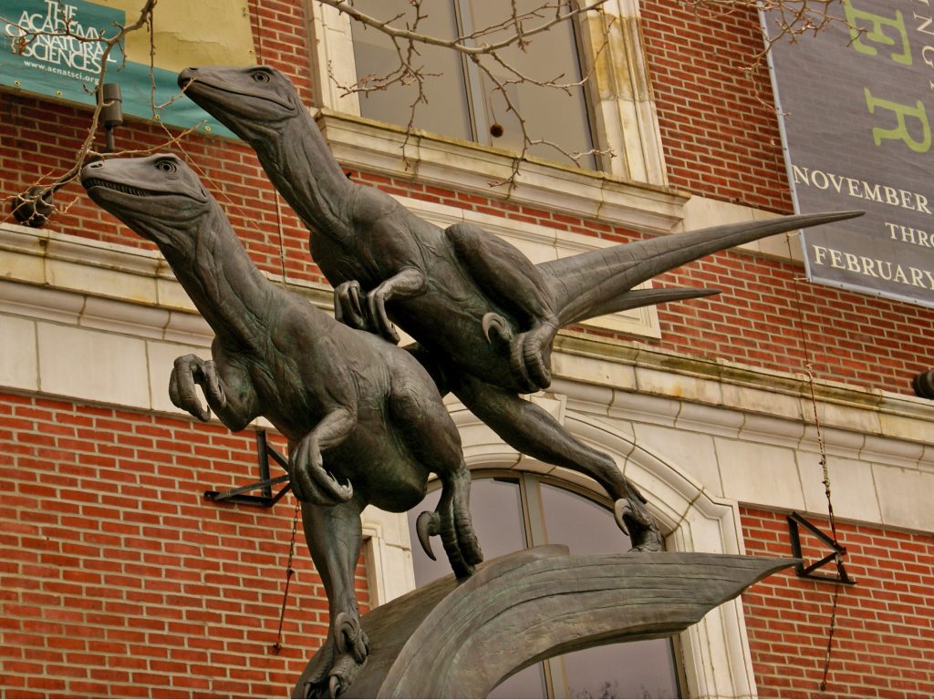 Two dark, metallic dinosaur sculptures, resembling raptors, are depicted in dynamic, leaping poses. One is positioned slightly above the other, both looking upwards and to the left against a brick building.