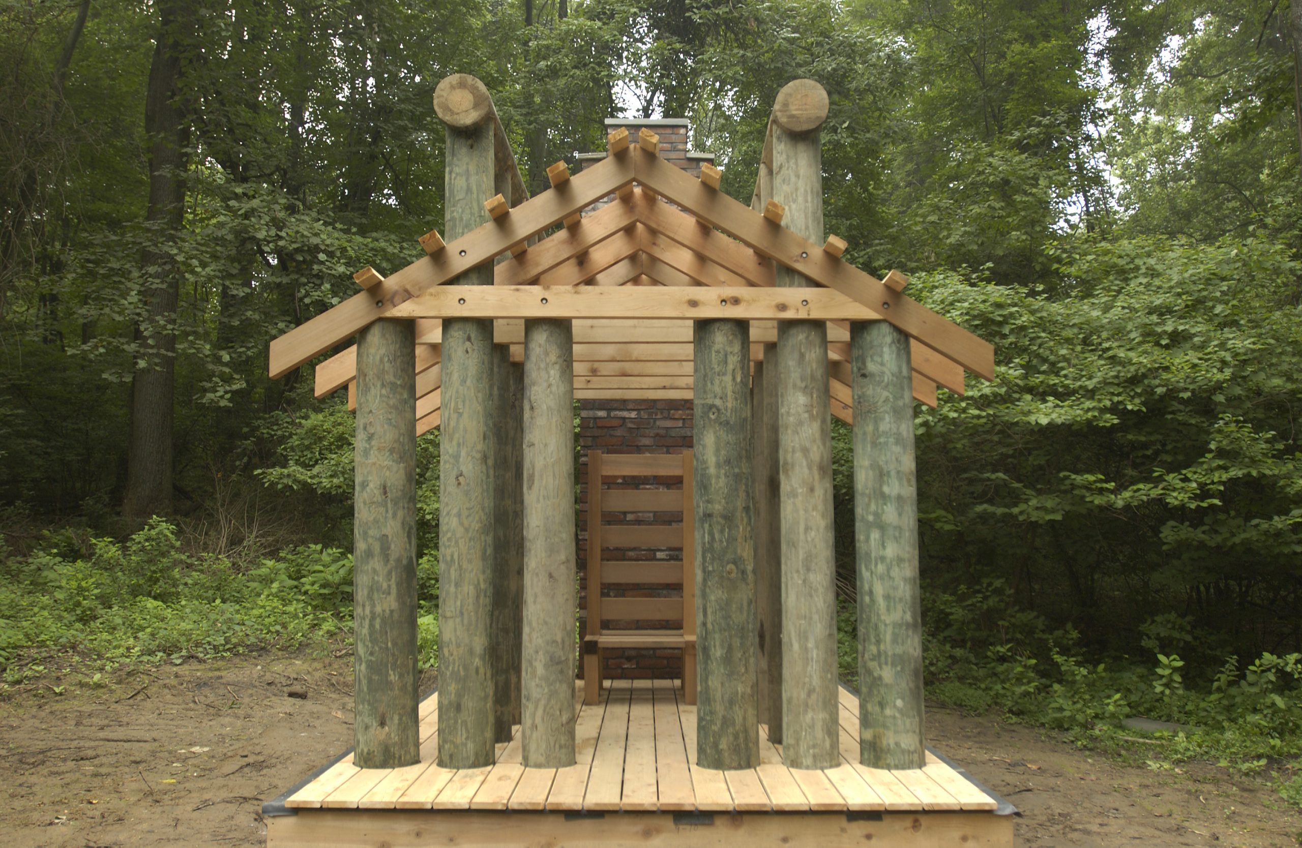 A symmetrical, front-facing view of a log-cabin architectural sculpture with a wooden chair inside, set in a forest clearing.