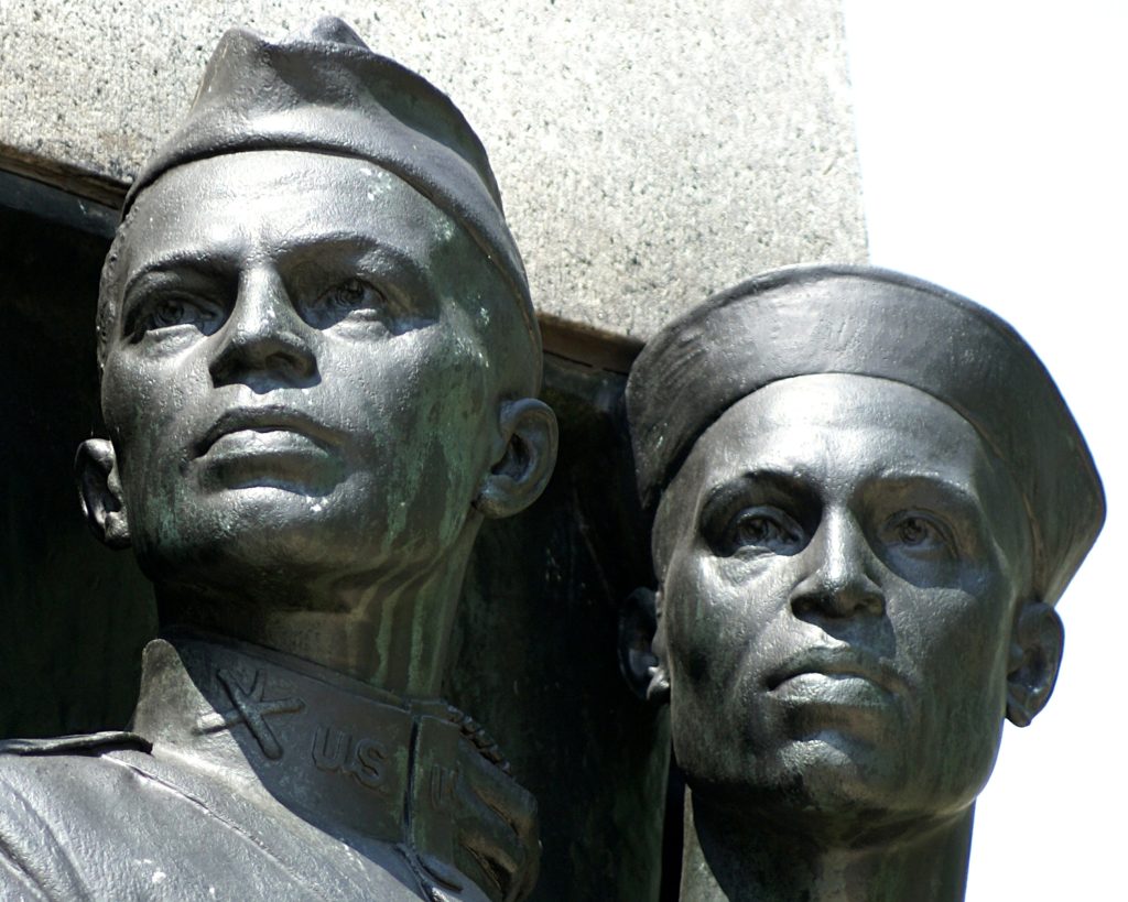 Two bronze busts of men in military uniforms. The figure on the left wears a military cap and a uniform collar with 'US' and an insignia. The figure on the right wears a sailor's cap. Both look slightly upwards.