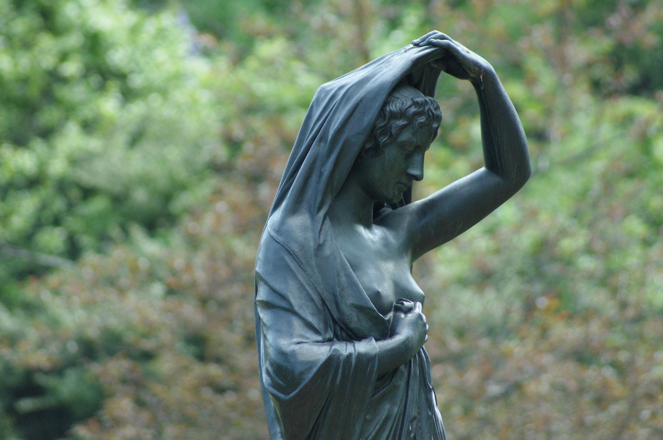 A medium close-up shot of Edward Stauch's bronze sculpture "Night," portraying a classical draped woman drawing a veil over her head against a blurred green backdrop.