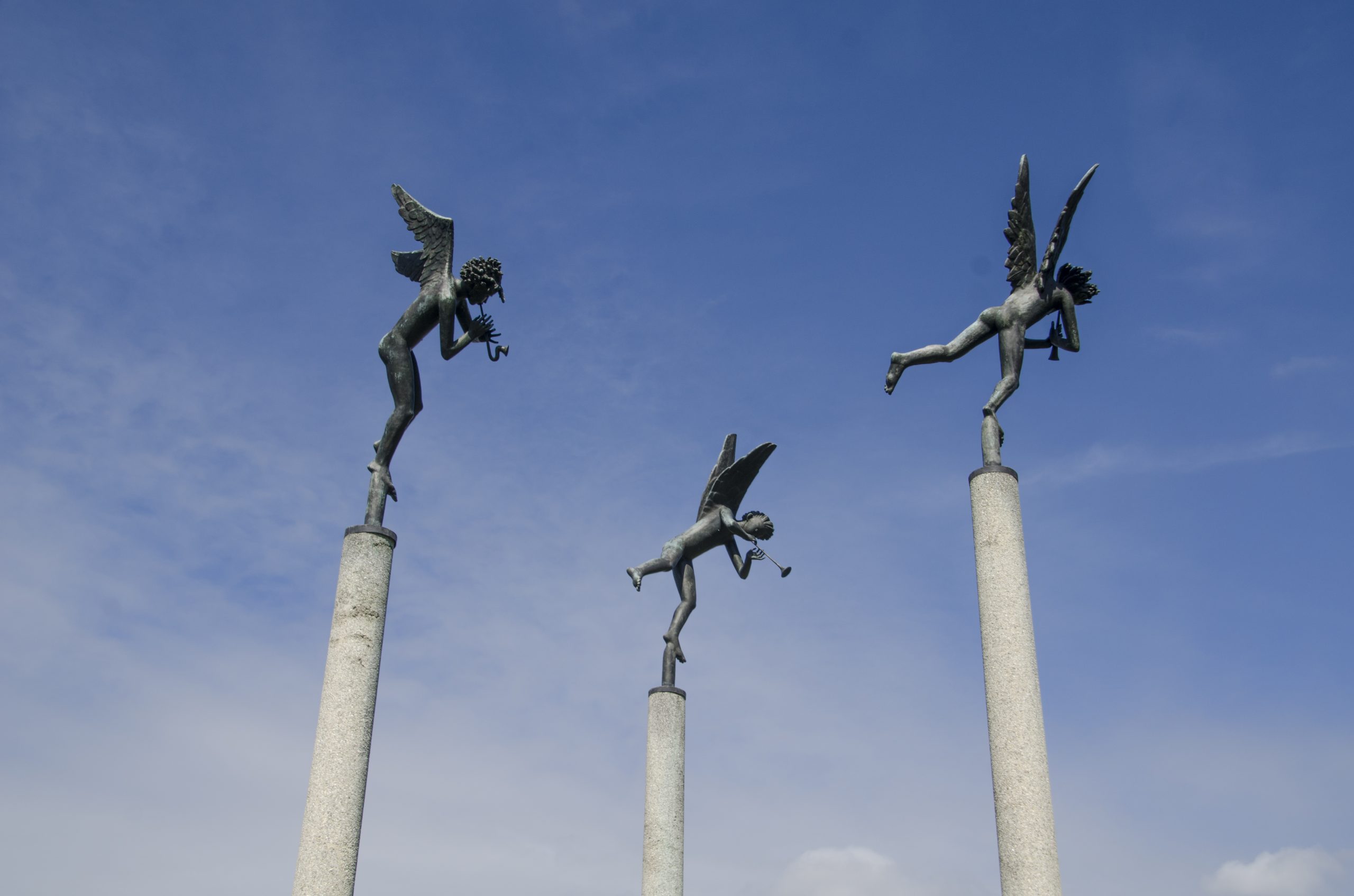 A low-angle view of Carl Milles' "Playing Angels" sculpture, showing three bronze angel figures on tall concrete pillars playing horns against a blue sky.