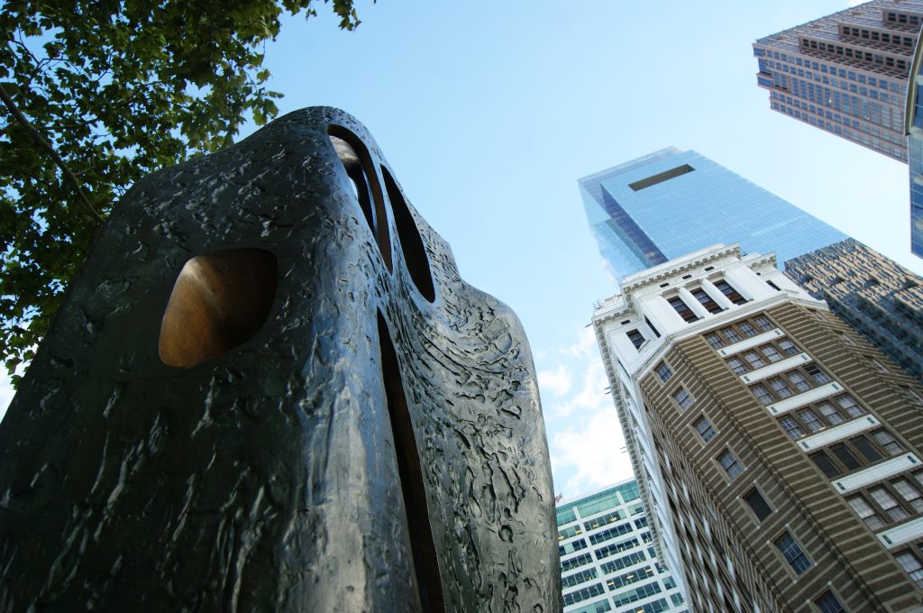 A large, dark, textured sculpture with organic, rock-like forms and openings dominates the foreground, viewed from a low angle. Behind it, tall modern glass skyscrapers and an older building rise against a clear blue sky, with a tree on the left.
