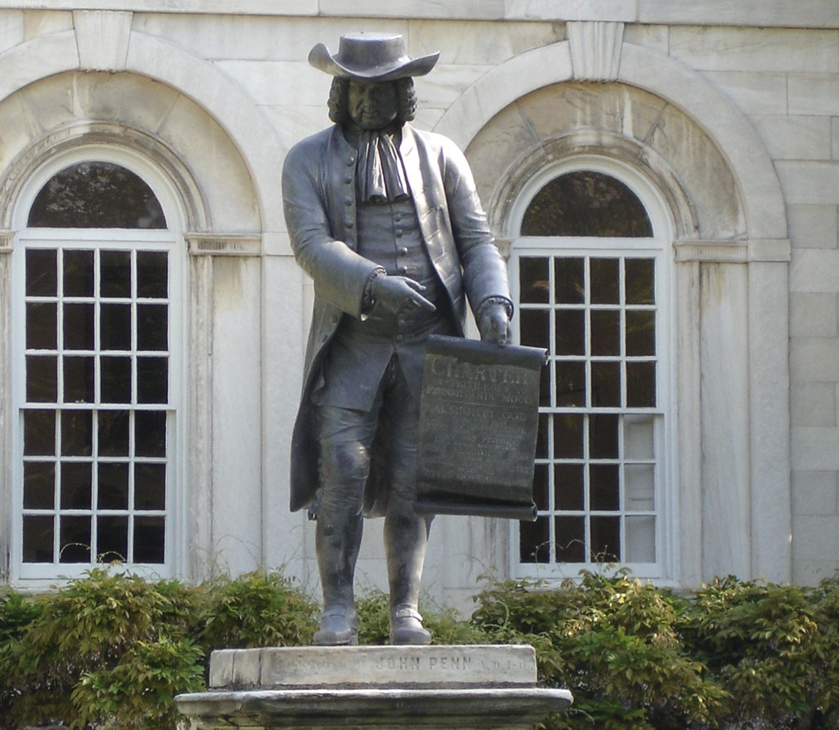 A full-length bronze statue of a man in 18th-century colonial attire, complete with a tricorn hat, holding and pointing to an unrolled scroll document labeled "CHARTER" in front of a white building with arched windows.