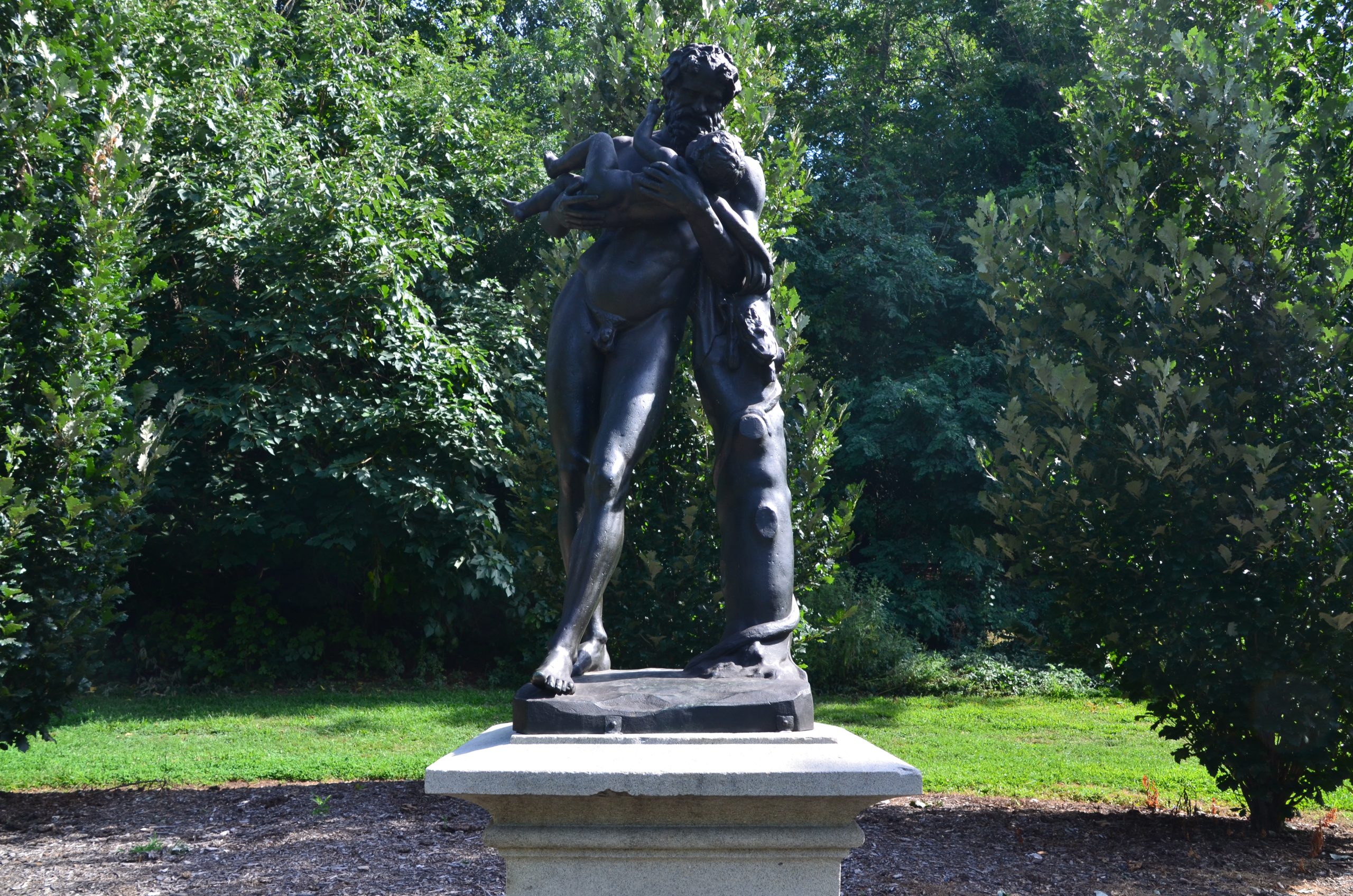 A bronze sculpture depicts a muscular, bearded man holding a partially draped baby. The man is nude and stands on a light-colored pedestal against a background of green trees.