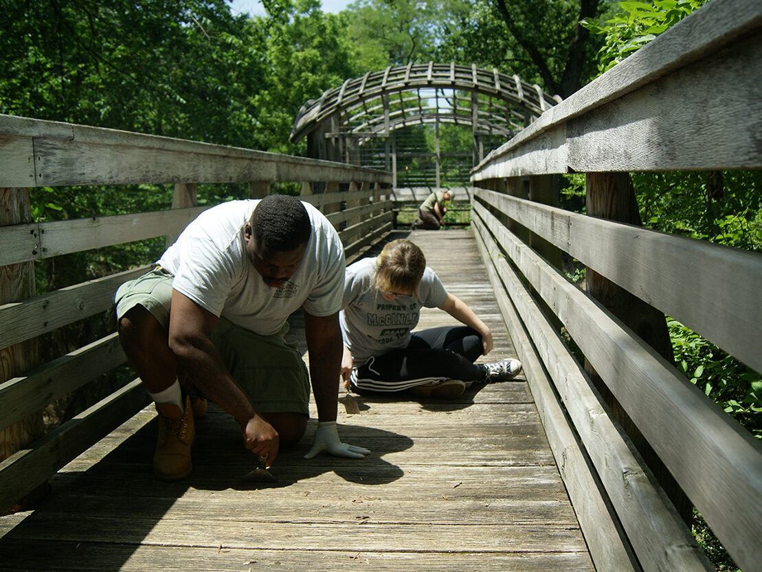 Two conservators performing work on the wooden walkway that leads to the treehouse-like "Pavilion in the Trees" observation platform in Fairmount Park