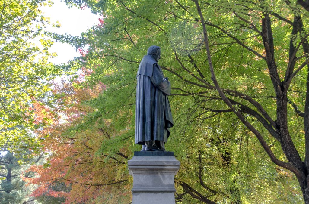 A bronze statue of a man in a long cloak stands on a stone pedestal, surrounded by trees with green and autumn-colored leaves.