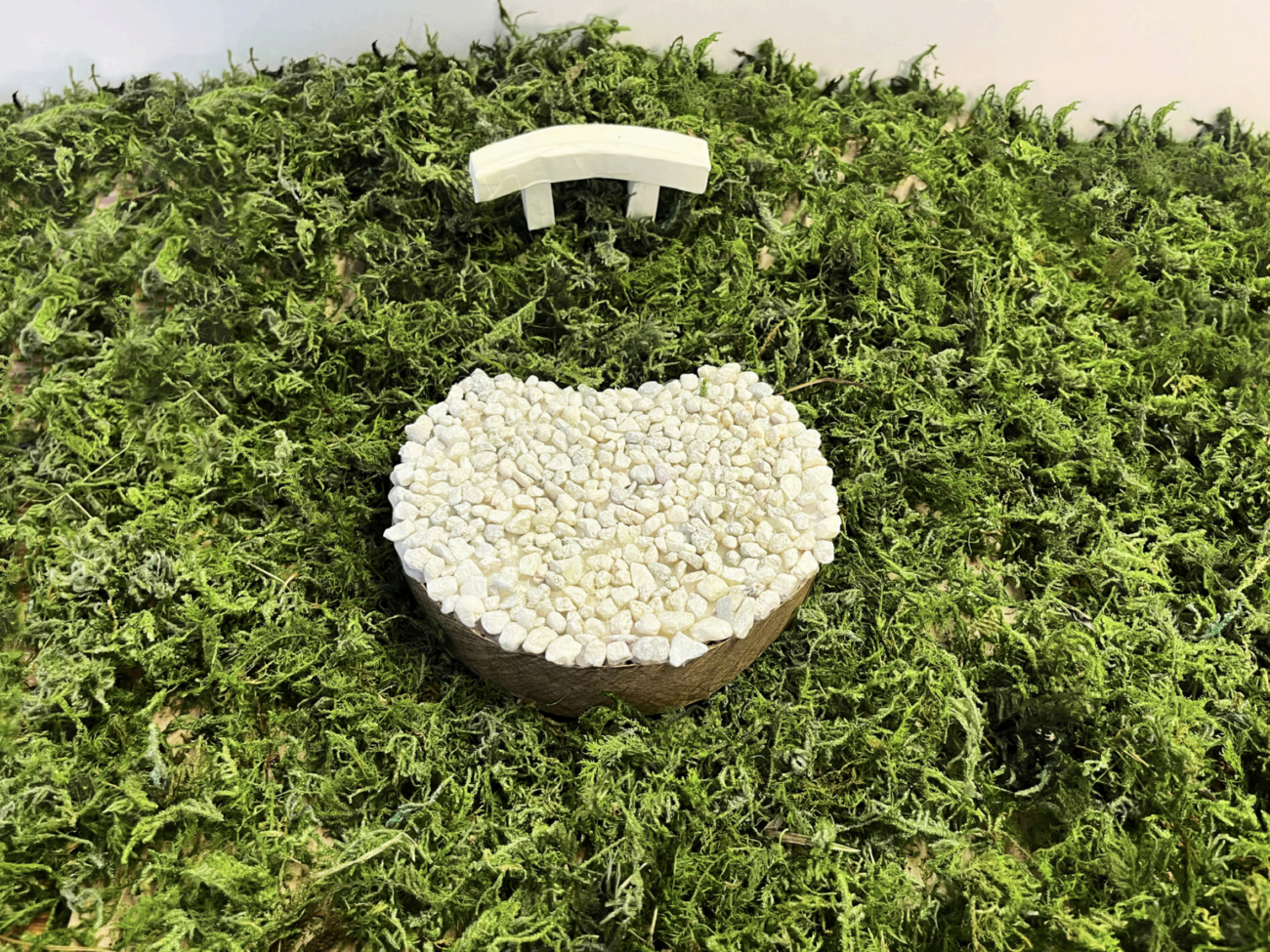 Looking down at the maquette of an artist's proposed installation. The artwork looks like a circular platform in the grass, depicted here with mossy material, embedded with light-colored stones. A light colored bench sits next to the circular platform.