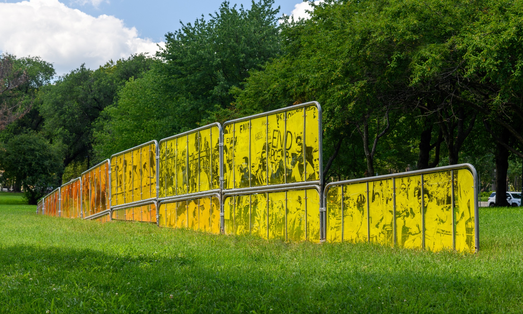 An outdoor art installation features a series of metal-framed panels with translucent yellow and orange inserts, arranged in a line on a grassy lawn. The panels display black silhouettes of people and text. Trees are visible in the background under a blue sky.