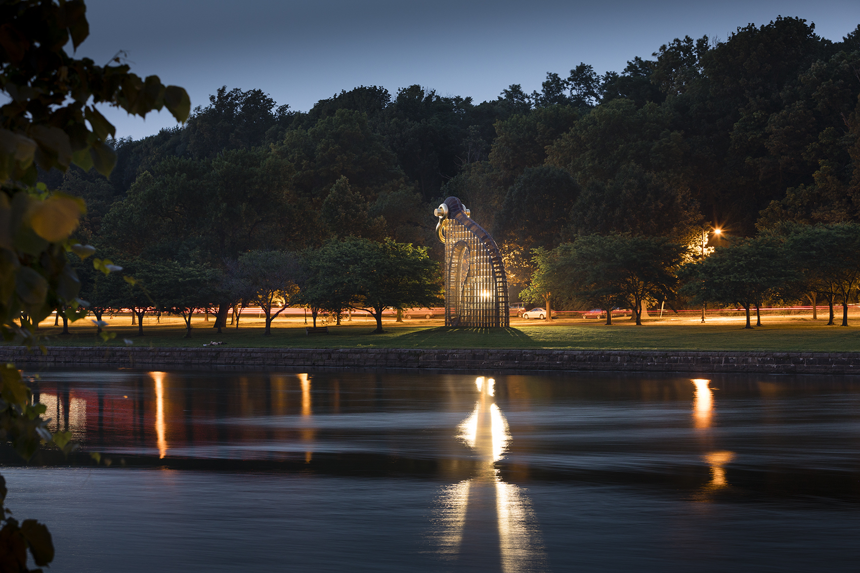 A large, illuminated, cage-like sculpture of a kneeling human figure stands in a grassy park at dusk. Behind it, a road with streaks of car lights and streetlights runs alongside a dense treeline. In the foreground, a body of water reflects the lights.