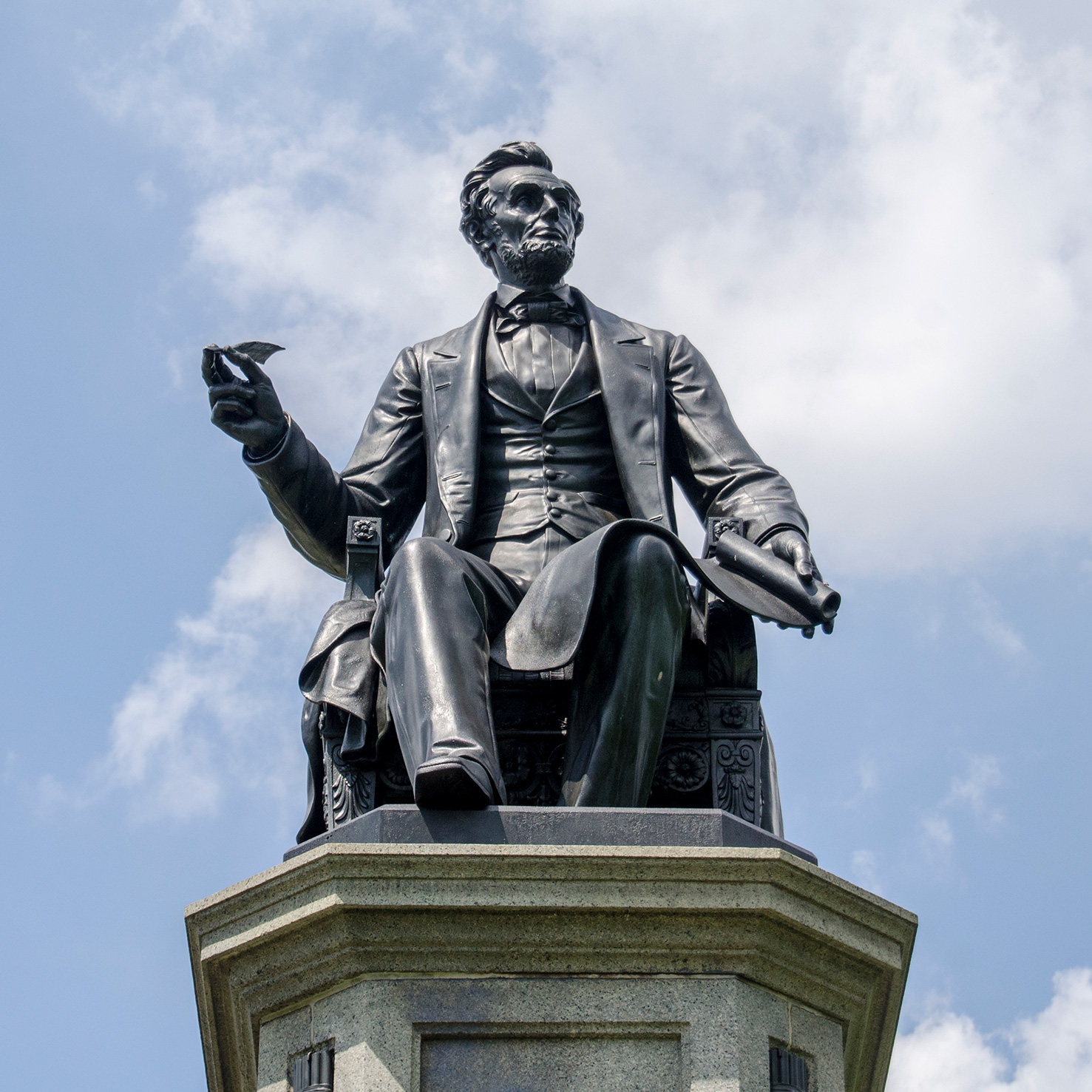 Bronze statue of a seated Abraham Lincoln with quill in hand against a blue sky with clouds.