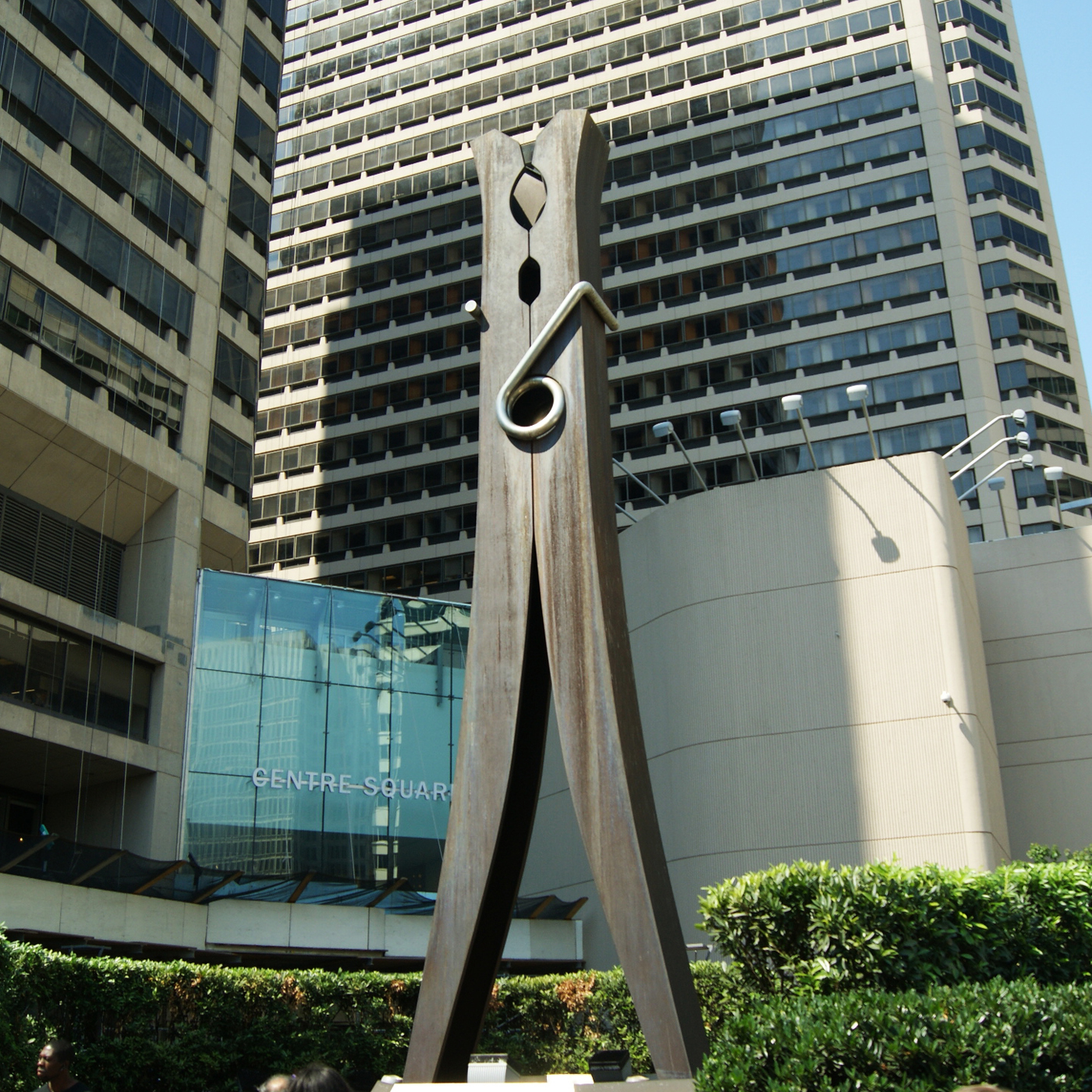 A monumental, dark metal sculpture resembling a clothespin stands upright in an urban setting. It features a large circular spring mechanism and is set amidst green hedges, with tall, modern buildings in the background.