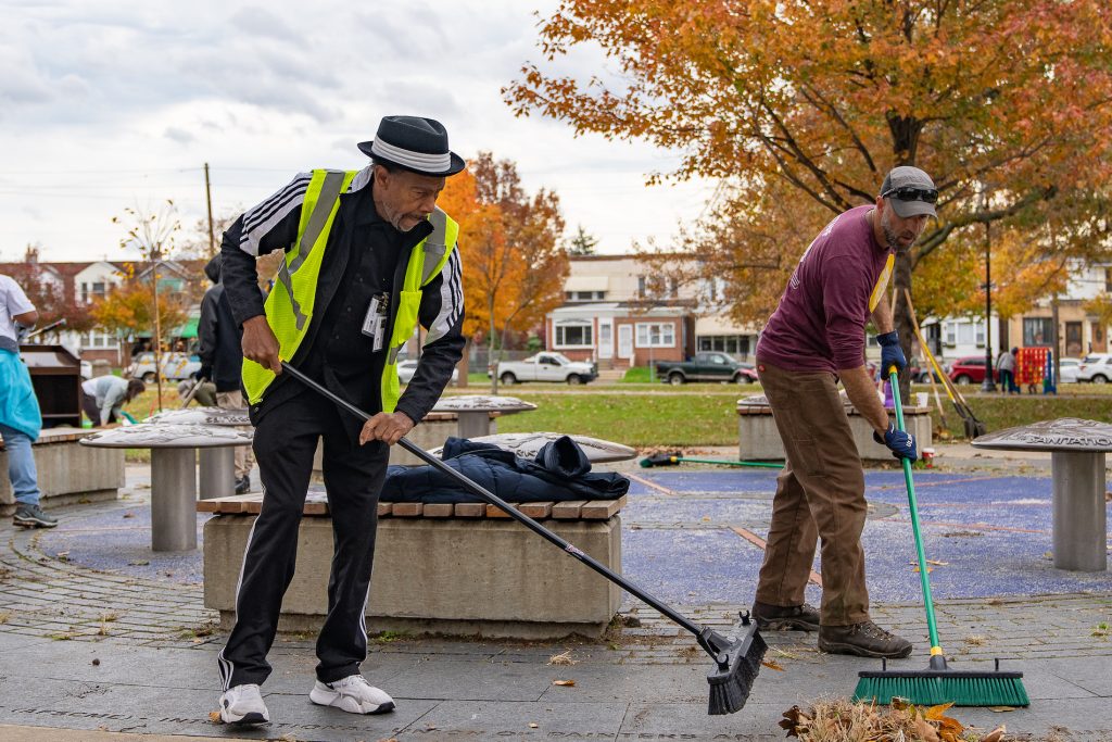 two men participating in the City's "Love Your Park" cleanup event, sweeping leaves at The Labor Monument in Southwest Philadelphia