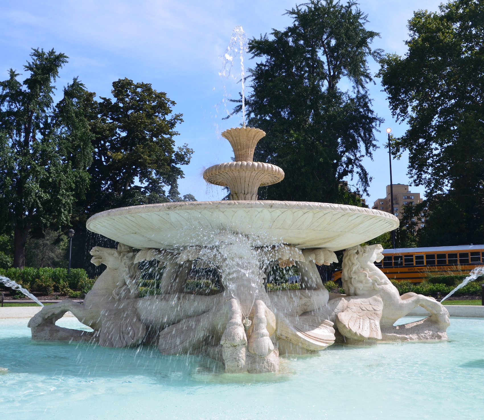 A large, multi-tiered stone fountain with water cascading from a wide basin supported by sculpted seahorse figures. The fountain sits in a pool of light blue water with green trees in the background.
