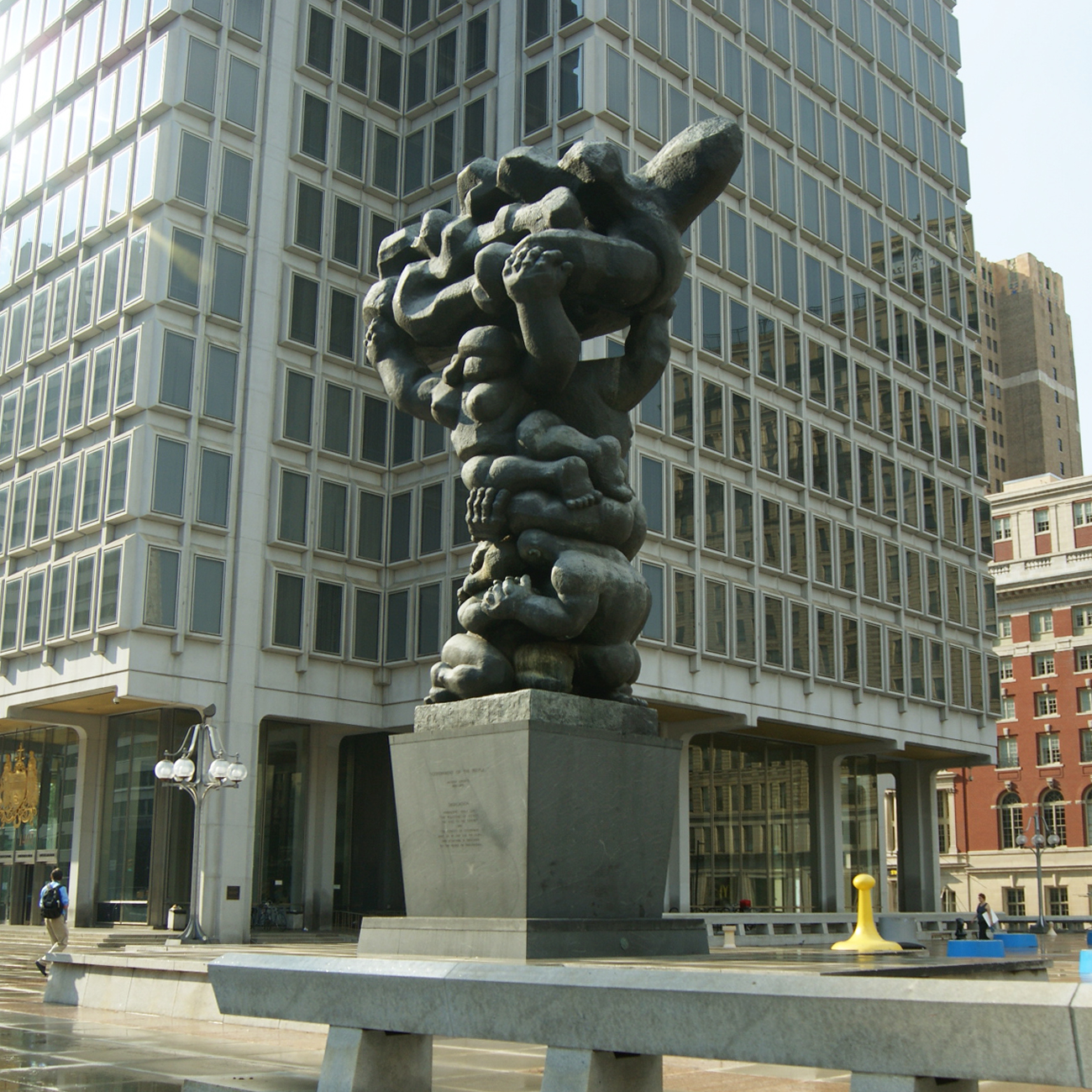 A large, dark bronze sculpture of numerous intertwined human figures forming a towering mass, set on a tall gray pedestal. It stands in front of a modern glass-faced building in an urban plaza.