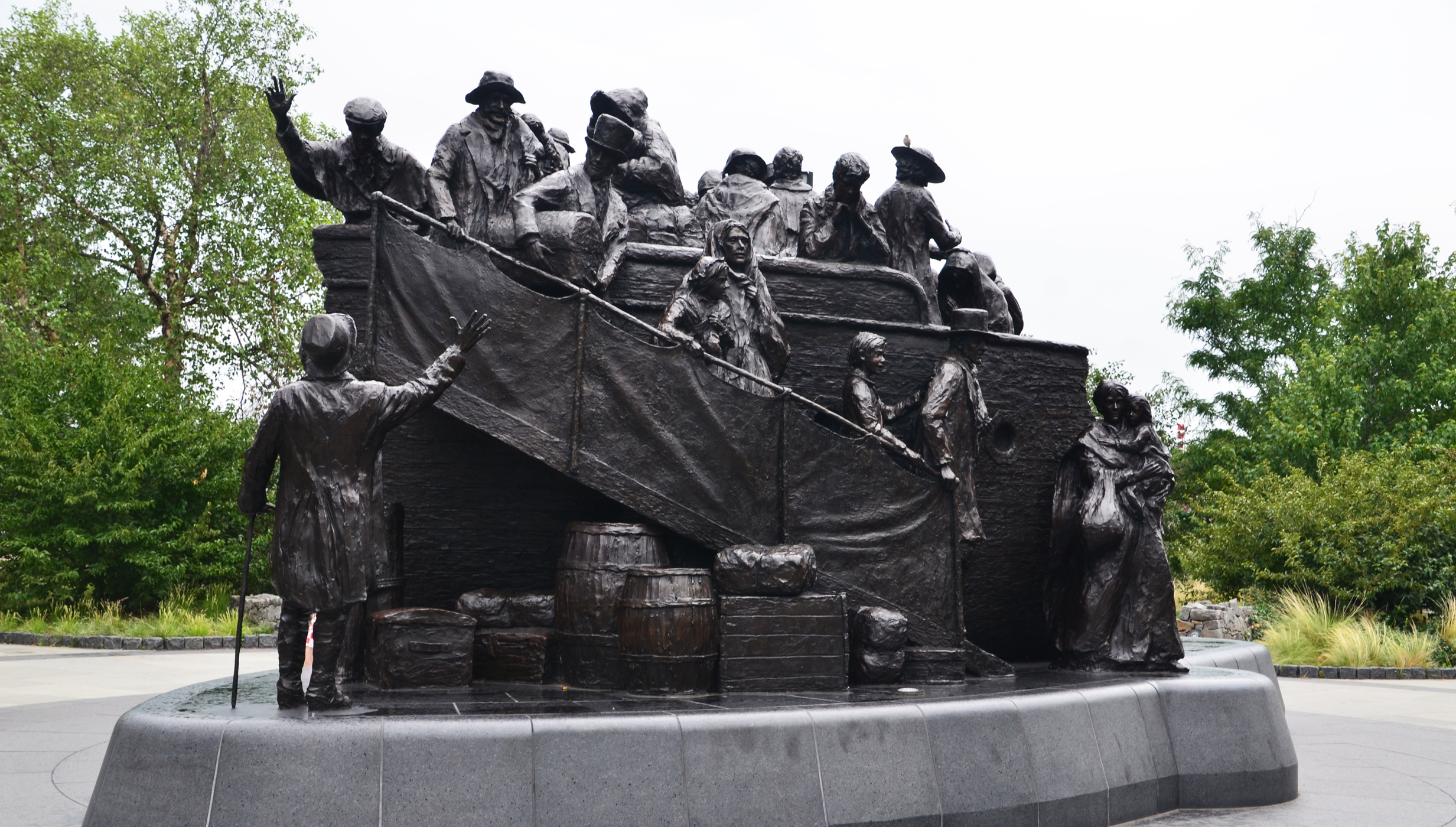 A large, dark bronze sculpture shows a group of gaunt figures in period clothing, some on a boat-like structure, others standing on the ground. Many figures carry bundles, and one holds a child, conveying a scene of hardship and migration. The sculpture is set on a dark, tiered base outdoors.