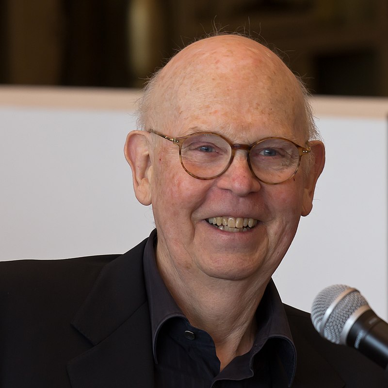 Portrait of artist Claes Oldenburg. He is older, bald, smiling, and wearing glasses. He has a microphone in front of him.