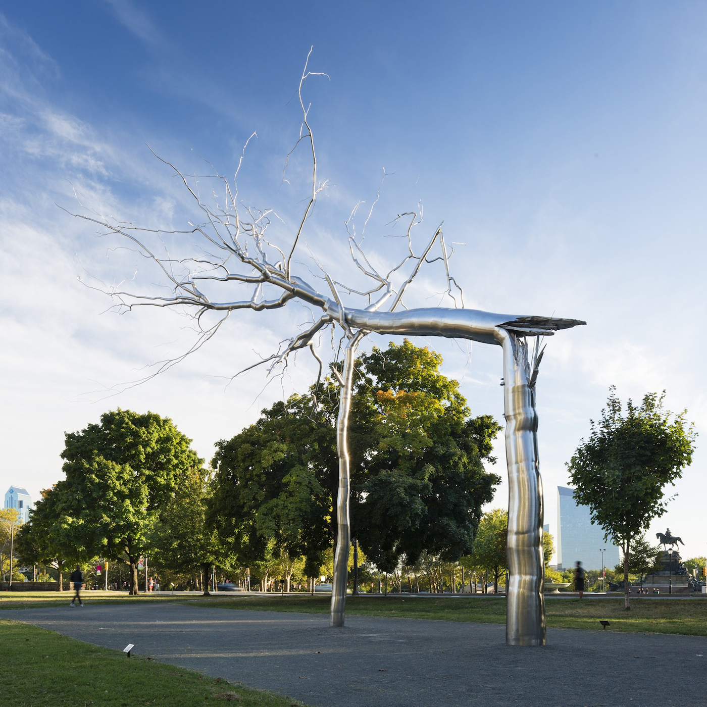 A large, polished metal sculpture of a tree with long, horizontal, wind-swept branches stands in a park. Green trees, a paved path, and a blue sky are in the background.