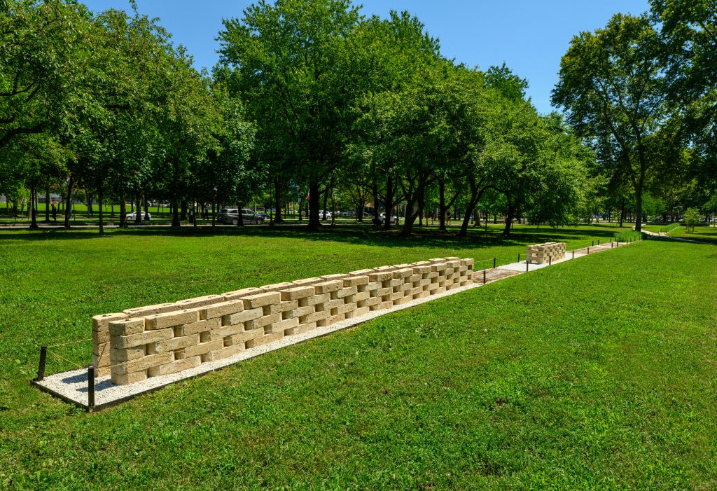 A long, low linear sculpture made of stacked light-colored rectangular blocks on a gravel path. The sculpture extends through a green grassy park with numerous mature trees, with a street and cars visible in the distance.