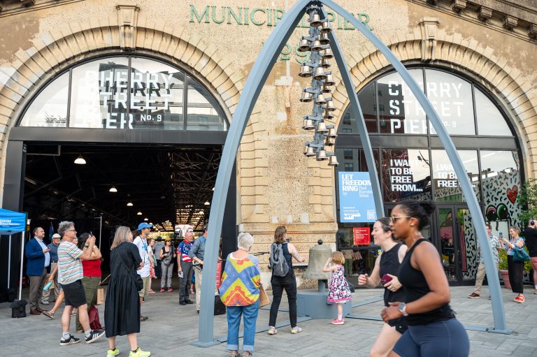 A public art installation featuring a tall column of small bells suspended between massive curved metal arches stands in front of the historic stone facade of Cherry Street Pier, while pedestrians and a jogger pass by.