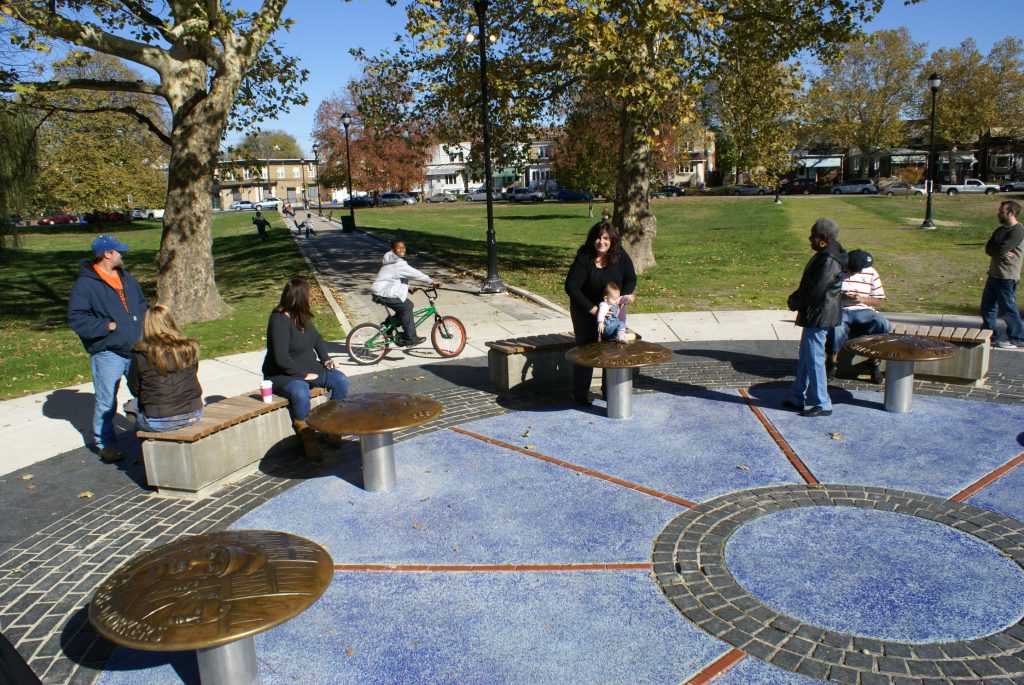 A public park scene features a circular blue ground mosaic surrounded by bronze discs on pedestals and benches. Several people are present, including a woman holding a baby near the installation and a person riding a bicycle on a paved path.