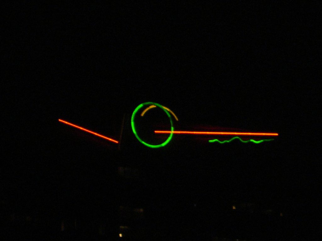A dark image features neon lights. A bright green circle is central, with a short, curved orange line inside it. To the left is a diagonal orange line, and to the right, a horizontal orange line with a wavy green line below it.
